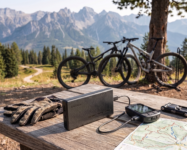 Nitecore NB10000 Gen 4 carbon fiber power bank charging a GPS bike computer on a wooden table with mountain bikes and alpine peaks in the background